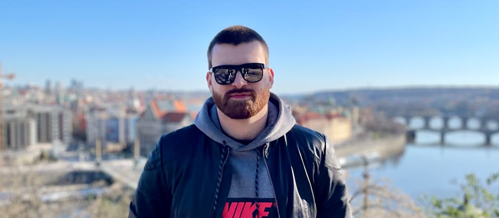 Aleksandr Gumroian standing in sunglasses, the background is out of focus with a cityscape, a river and bridges.